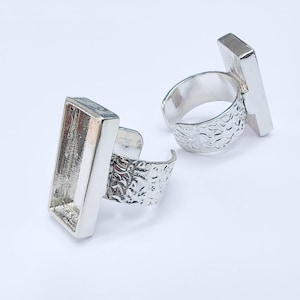 Może przedstawiać: Dwa srebrne regulowane pierścionki z prostokątnymi oprawami. Paski mają wzór tekstury, a oprawy są puste, gotowe do personalizacji. Pierścionki są prezentowane na białym tle.