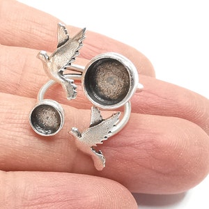 Könnte beinhalten: Ein silberfarbener Ring mit einem Taubendesign. Der Ring zeigt zwei fliegende Tauben mit zwei kreisförmigen Fassungen für Verzierungen. Der Ring wird auf einer Hand vor weißem Hintergrund gezeigt.