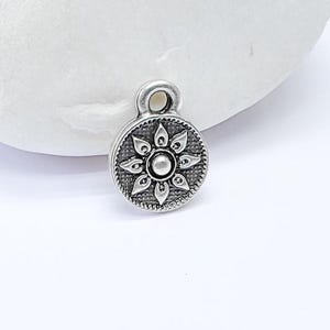 May include: Silver charm with a detailed floral design, resembling a flower with a central circle. The charm has a loop at the top for attaching to jewelry.