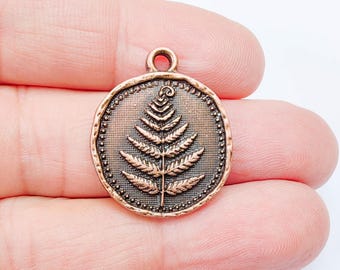 May include: A round, copper-colored pendant with a detailed fern leaf design. The pendant has a textured border with small, raised dots and a loop at the top for attaching to a chain or cord. The fern is centered on a textured background.