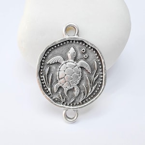 May include: A silver-toned metal charm featuring a detailed turtle design. The charm has a circular shape with a textured border and two loops for attachment. The turtle is surrounded by aquatic plants and bubbles.