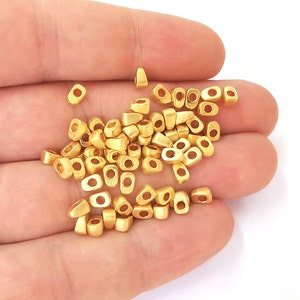 May include: A collection of small, matte gold-colored beads. Each bead has a rectangular shape with rounded edges and a central hole. The beads are scattered, suggesting they are loose and ready for use in jewelry making or crafts.