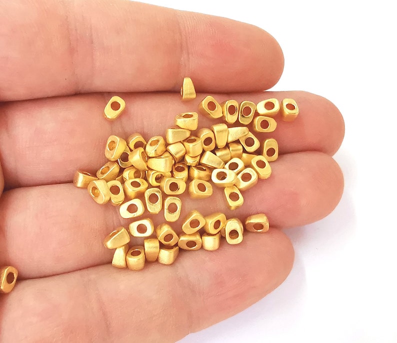 May include: A collection of small, matte gold-colored beads. Each bead has a rectangular shape with rounded edges and a central hole. The beads are scattered, suggesting they are loose and ready for use in jewelry making or crafts.