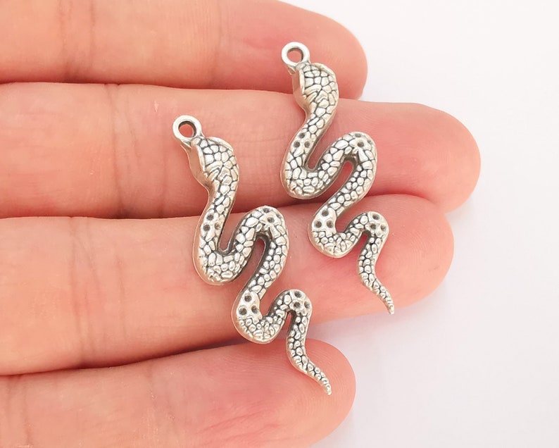 May include: Two silver snake charms with a textured surface. The charms are in the shape of a snake with its head raised and its body curled.