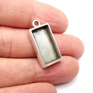 Pode incluir: Pingente retangular prateado com um inserto de vidro transparente. O pingente tem um laço na parte superior para prender a uma corrente ou cordão.