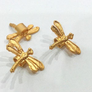 Puede incluir: Dos abalorios de libélula de color dorado con detalles intrincados. Los abalorios están diseñados para ser utilizados como hallazgos de joyería o adornos.