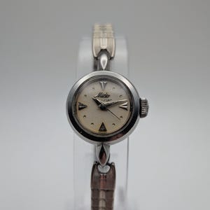 Op de afbeelding: Een vintage Mido dames polshorloge met een zilverkleurige metalen band en een ronde wijzerplaat. De wijzerplaat is gebroken wit met zilveren wijzers en uurmarkeringen. Het horloge wordt op een transparante standaard getoond.