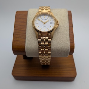 Könnte beinhalten: Goldfarbene Pulsar-Uhr mit weißem Zifferblatt, Datumsanzeige und goldenem Gliederarmband. Die Uhr wird auf einem Holzständer mit einem beigen Kissen präsentiert. Auf dem Zifferblatt steht das Wort "Pulsar".