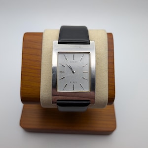 May include: A rectangular Guess watch with a silver face and a black leather strap. The watch face has silver hands and markers. The word "STEEL" is printed on the face. The watch is displayed on a wooden stand.