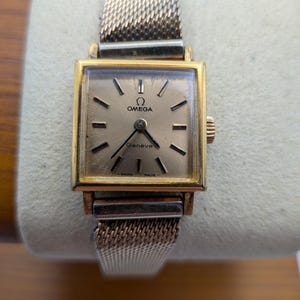 May include: A vintage Omega Genève wristwatch with a gold-toned square face and a mesh band. The watch face is a light gold color with black hands and markers. The watch is displayed on a cream-colored surface.