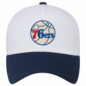 Könnte beinhalten: Weißes Baseball-Cap mit marineblauem Schirm. Die Kappe hat ein gesticktes Logo eines Basketballs mit dem Text "76ers" in Rot und Blau. Die Kappe hat einen gebogenen Schirm und einen Knopf oben.