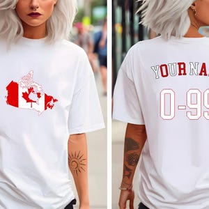 Puede incluir: Camiseta blanca con un gráfico rojo y blanco de la bandera canadiense. La parte trasera de la camiseta muestra el texto "YOUR NAME" sobre el número "0-99" en rojo y blanco.