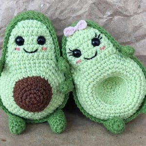 Puede incluir: Dos peluches de aguacate tejidos a crochet con caras sonrientes. Un aguacate tiene un lazo rosa en la cabeza.