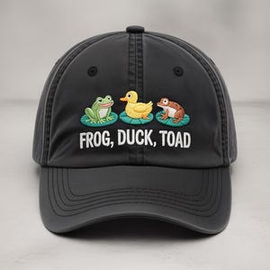 Pode incluir: Boné de beisebol cinza escuro com imagens bordadas de um sapo verde, um pato amarelo e um sapo marrom. O texto "FROG, DUCK, TOAD" é bordado abaixo. O boné tem uma aba curva.