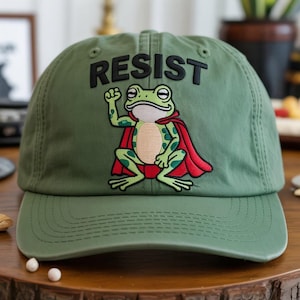 Puede incluir: Gorra de béisbol verde oliva con la palabra "RESIST" bordada en negro. La gorra presenta una rana bordada con una capa roja. La rana es verde con manchas amarillas y está haciendo un puño.