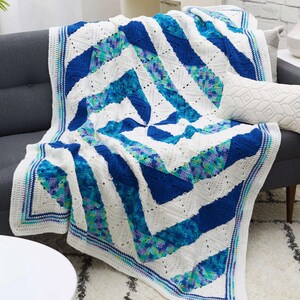 Pode incluir: Uma manta de crochê branca com padrões geométricos azuis e verdes. A manta está colocada sobre um sofá cinza com uma almofada branca.