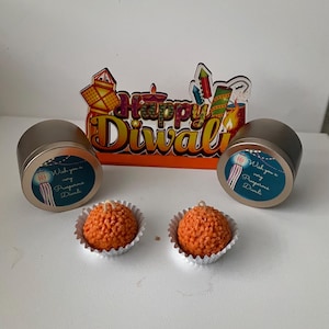 May include: Two round, orange candles in silver foil cups, next to two silver tins with teal labels. The labels read "Wish you a very Prosperous Diwali" with a lantern graphic. A decorative sign reads "Happy Diwali".