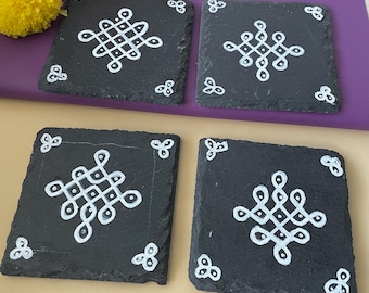 May include: Four square black slate coasters with a white geometric design in the center and white corner accents. The coasters are arranged on a purple and tan surface, with orange and yellow flowers in the background.