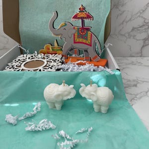 May include: An open box with a teal interior holds decorative items. Two white elephant candles sit in front of a wooden elephant cutout with a decorative umbrella. A decorative wooden frame and other cutouts are also in the box.