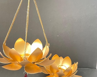May include: Two gold-coloured lotus flower candle holders. One is suspended by a gold chain, and the other rests on a surface. A red crystal hangs from the suspended holder. The candle holders are designed to hold tea light candles.