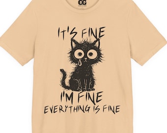 Op de afbeelding: Beige t-shirt met een zwarte illustratie van een verdrietige kat met grote ogen en de tekst "IT'S FINE I'M FINE EVERYTHING IS FINE" in handschrift. Het shirt heeft korte mouwen en een ronde hals.