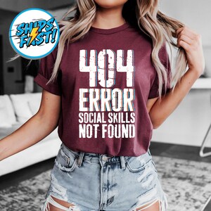 May include: Burgundy t-shirt with the text "404 ERROR SOCIAL SKILLS NOT FOUND" in white, with a blue and red shadow effect. The shirt has short sleeves and is worn with denim shorts.