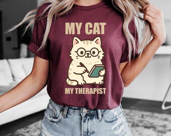 May include: Burgundy t-shirt with the text "MY CAT MY THERAPIST" above a cartoon cat wearing glasses and holding a notepad. The cat is beige with black stripes and is drawn in a cute style.