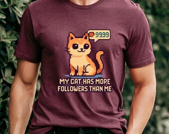 May include: Burgundy t-shirt featuring a pixelated orange cat with a speech bubble displaying "9999". The text below the cat reads "MY CAT HAS MORE FOLLOWERS THAN ME".