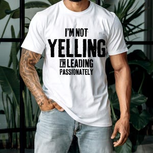 May include: White t-shirt with the text "I'M NOT YELLING I'M LEADING PASSIONATELY" in bold black letters. The shirt is a classic crew neck style and is being worn by a person. The person is wearing light blue jeans.