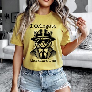 May include: Yellow t-shirt with the text "I delegate therefore I am." The shirt features a black and white illustration of a cat wearing a hat, sunglasses, and a suit. The cat's face is centered on the shirt.