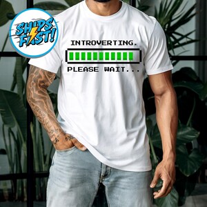 May include: White t-shirt with the text "INTROVERTING. PLEASE WAIT..." above a green loading bar graphic. The shirt is a crew neck style and is being worn.