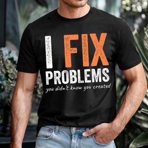 Puede incluir: Camiseta negra con el texto "I FIX PROBLEMS you didn't know you created" en letras blancas y naranjas. La camiseta es de cuello redondo y se está usando.