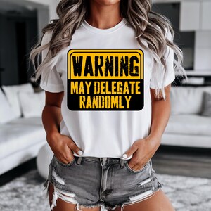 May include: White t-shirt with a black and yellow warning sign graphic. The sign reads "WARNING MAY DELEGATE RANDOMLY" in bold, sans-serif font. The shirt is worn with gray denim shorts.