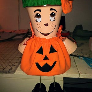 May include: A whimsical, hand-painted terracotta flower pot transformed into a smiling jack-o'-lantern figure. The pot features a green brim, an orange pumpkin costume, and black painted feet. The face has painted eyes, freckles, and a heart-shaped nose.
