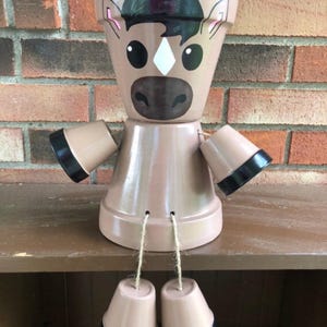 May include: A whimsical, handcrafted horse-shaped flower pot made from terracotta. The pot features painted details including a black mane, brown muzzle, and white diamond-shaped marking. The arms and legs are attached with twine, and the hooves are painted black.