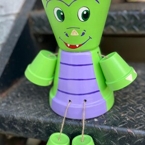 May include: A whimsical, hand-painted dragon-shaped flower pot. The pot is primarily lime green with purple and white accents. The dragon has large green eyes, a smiling mouth, and small pot arms and feet. The pot is sitting on a metal surface.