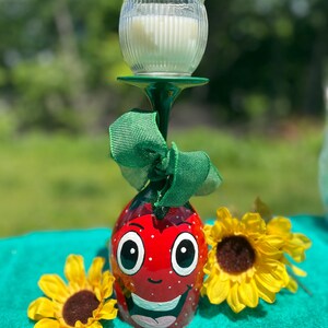 May include: A red wine glass painted to look like a smiling strawberry with white dots. The glass is holding a clear glass candle holder with a white candle inside. The glass is decorated with a green ribbon and is sitting on a turquoise surface with two yellow sunflowers.
