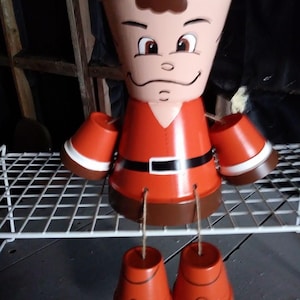 May include: A whimsical, hand-painted terracotta pot figure. The figure is painted with a face, a red outfit, and brown accents. The arms and legs are attached with twine, and the figure is standing on a white wire shelf.