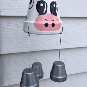 May include: A handmade cow-themed wind chime. The top pot is painted white with a cow face design, featuring black eyes, ears, and a pink snout. Three silver-painted pots hang below. The wind chime is suspended by a pink string.