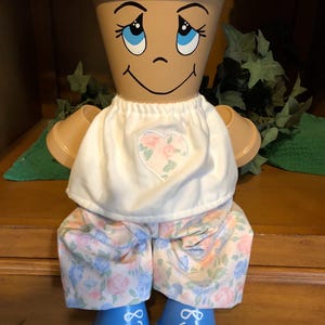 May include: A whimsical, hand-painted terracotta flower pot doll. The doll has a smiling face with blue eyes, wearing a white top with a heart-shaped floral design, and floral patterned pants. The doll's feet are painted blue with white accents.