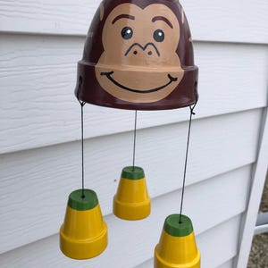May include: A wind chime made from terracotta pots painted to look like a monkey face with a smiling expression. The monkey face pot is brown with a tan face and black eyes. Three smaller yellow pots with green tops hang below the monkey face pot.