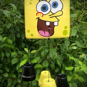May include: A yellow wind chime with a painted image of Spongebob Squarepants. The chime has four hanging bells, two black and two yellow.