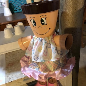 May include: A whimsical, handmade terracotta flower pot doll. The doll has a painted face, arms, and legs made from smaller pots. It wears a dress with a white top and a pink skirt. The doll is a unique decorative item.