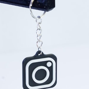 May include: A black and white Instagram logo keychain. The keychain features the iconic camera logo with a white outline and a silver chain. The keychain is hanging against a white background.