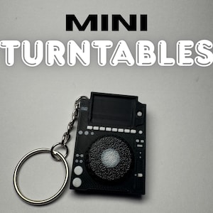 Peut inclure: Porte-clés mini platine vinyle noir avec un anneau argenté. Le porte-clés présente un design détaillé, comprenant un cadran central et divers boutons. Les mots "MINI TURNTABLES" sont affichés en lettres blanches au-dessus.