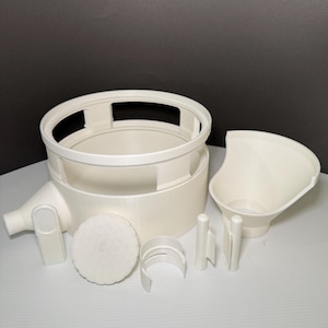 May include: A collection of off-white kitchen tools, including a large circular strainer, a funnel, a filter, and several small support pieces. The items are arranged on a white surface against a dark gray background.