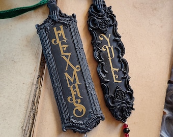 May include: Two black decorative bookmarks with gold lettering and ornate frames. One reads "HEXMAS" and the other "YULE". Each has a velvet ribbon and beaded charm. The bookmarks rest on an open book.