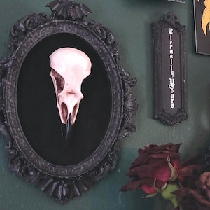 May include: A pale bird skull mounted inside an oval black frame with ornate detailing and a bat motif at the top. Next to it is a rectangular black plaque with the text Eternally Yours in white gothic lettering, and dark red roses below.