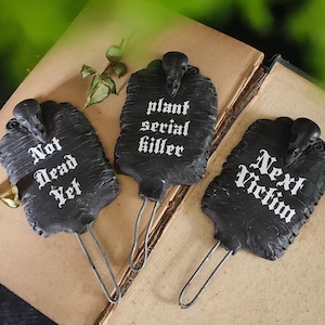 May include: Three black plant markers with bird skull toppers and metal stems. The markers have white text that reads "Not Dead Yet", "plant serial killer", and "Next Victim". The markers are on a book.