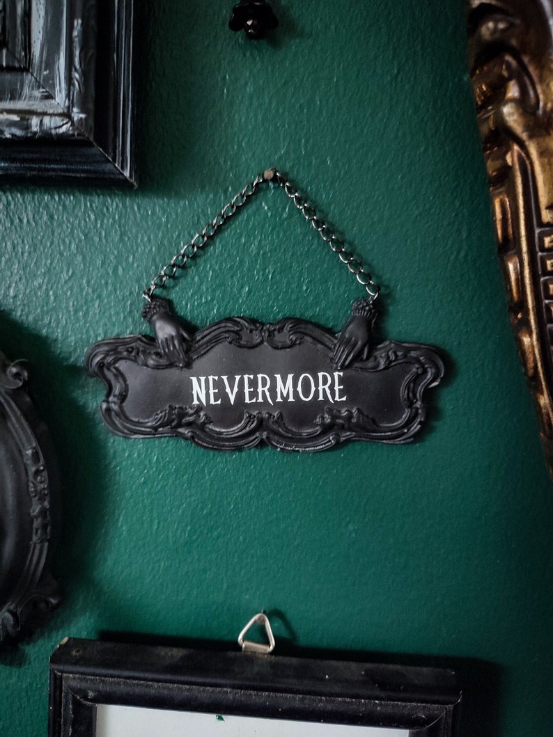 May include: A black metal sign with ornate detailing hangs from a chain. The sign reads "NEVERMORE" in white lettering.